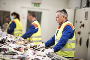Senior worker sorting papers on factory assembly line for recycling at recycling plant.